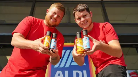 Jayden and Brendan Manders wear red t-shirts while each holding two bottles of sauce in front of a large Aldi sign. 