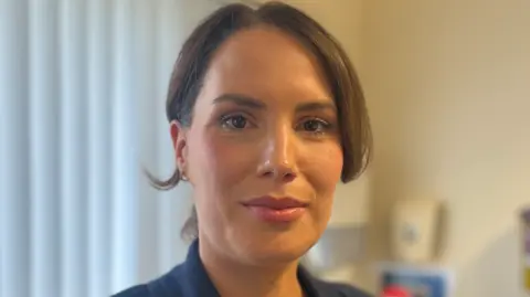 BBC Dr Jilly O'Hagan has brown hair parted in the middle and tied back in a pony-tail and has brown eyes. She is wearing a navy suit jacket