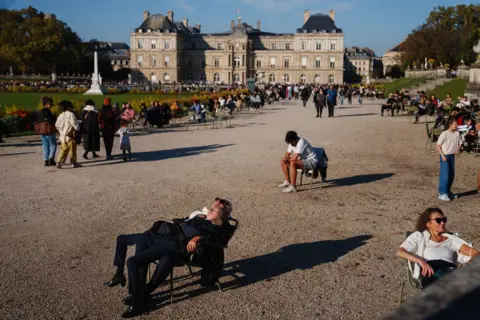 Dimitar DILKOFF AFP A woman lies on top of a man, both in black, lounging back on a chair in the foreground. One person in white sits on a chair behind them and a woman wearing white and sunglasses sits to the right. People walk along a promenade towards the Luxembourg Palace. Grass and trees line either site of the path.