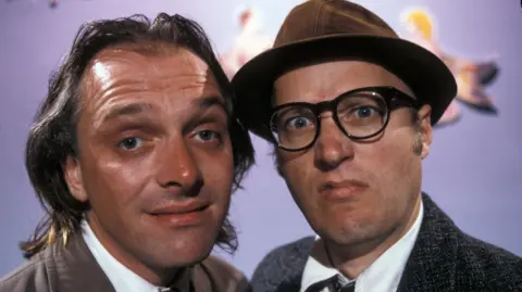 A promotional still for the sitcom Bottom, showing Rik Mayll with long, thinning, greasy hair, smiling effetely at the camera. He is standing next to Ade Edmondson, who is wearing glasses and a pork pie hat and glaring at the camera.