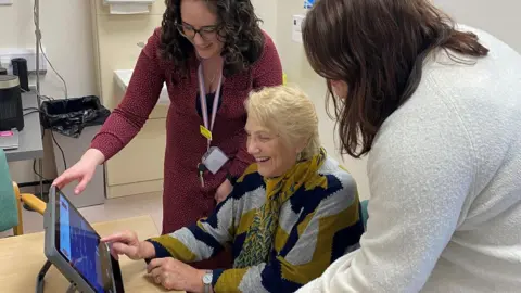 Two brown haired women stand around a woman with short blonde hair as she taps the screen of the device which looks like a large tablet.
