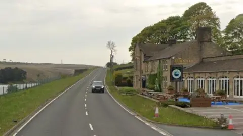 A car drives past the sandstone Turnpike pub on the A672, which lies alongside a grass verge and a reservoir, with moorland rising in the distance