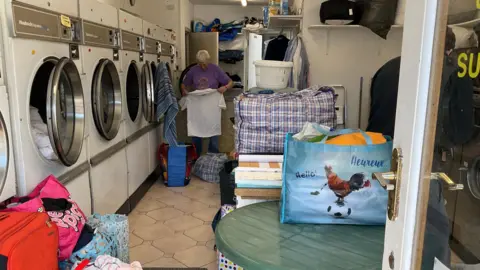 A picture of the inside of the launderette in Jersey. There is a number of washing machines pictured to the left, with bags of clothes surrounding it. There is a man in a purple shirt stood at the back of the room folding an item of clothing.