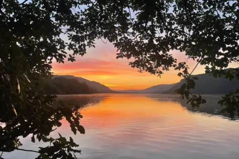 bbc weather watchers / Georgia Roper The sky is shades of yellow, orange, pink and purple behind the silhouettes of the rolling hills in the background. There is a ring of leafy branches in the foreground. The scene looks onto the lake beyond, which is so still the reflection of the mountains and the sky is cast in it.