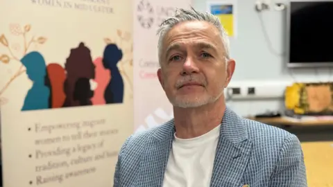 Michael McGoldrick is looking into the camera with a neutral expression. His short hair is white. He's wearing a white top with a blue and white checked jacket. A board is behind him. 