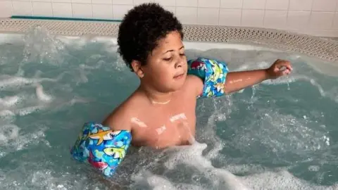 Family picture The picture shows a young child standing in a bubbling pool or hot tub filled with light blue, churning water and white foam. The child is chest-deep in the water and wearing inflatable arm bands with a bright, cartoon-style fish pattern in blue, green and yellow. One arm is extended out to the side just above the water, while the other is partly submerged.