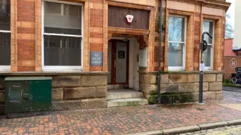 An orange-brick ornate old building with a cobbled pavement in front of it. A plaque reads: 161 High Street