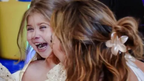 Family photograph Sienna, with long brown hair and wearing a sparkly cream and gold dress, laughs with her mum pictured from behind with long brown hair, tied back with a cream flower clip. They are hugging.