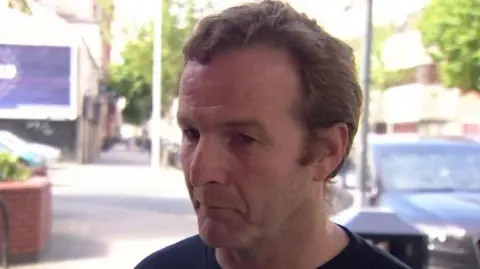 BBC A man in a navy top, with light brown hair, stands in front of a black car on a sunny day