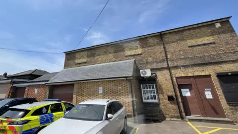 Dacorum Heritage Trust A brick building, with boarded up windows, a brown door, and one window. There is a garage shutter to the left and three cars parked outside, including a police vehicle. 