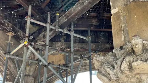 Inside the Poultry Cross. You can see scaffolding and the roof timbers which need repair, surrounded by the stone monument, which has arches and carving.