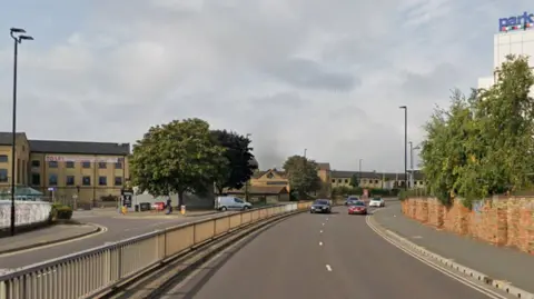 Google A Google Streetview image of Bourges Boulevard - a dual carriageway road with a metal barrier in the central reservation. The view is from the rear of a vehicle and there are cars driving up behind, and there is a van pulling out of a junction in the other carriageway. On the left there is a big three-storey brick building that says "to let" on the side, and there are some trees in full bloom. On the right is a brick wall next to a pavement that runs alongside the road. There are further trees and the edge of an industrial building behind the wall.