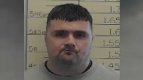 Grainy police mugshot of a man with short, cropped black hair. He is standing against a white height chart and staring directly at the camera with a serious expression on his face. He is wearing a grey sweatshirt with a black T-shirt underneath.