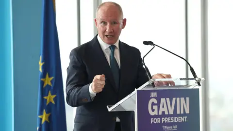 PA Media Jim Gavin, who is bald and wears a dark suit, heavy green tie and white shirt, stands behind a lectern. On it are the words 'Jim Gavin For President'. His mouth is open, his face is serious and he is gesturing with the knuckle of his index finger. Behind him is the European Union flag - a ring of yellows stars against a block of blue.