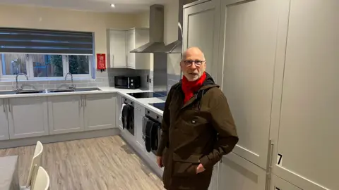 Vikki Irwin/BBC Richard O'Driscoll is standing in a kitchen of a new house, he is wearing a brown jacket and dark trousers. He has a red scarf wrapped around his neck. He is also waring glasses. 