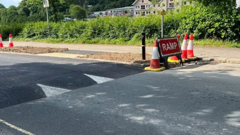 A table-top speed hump which forms part of a crossing installed on Malpas Road in Truro. Several traffic cones and a red warning sign saying ramp are on one side of the road. Buildings can be seen in the background past a hedgerow.