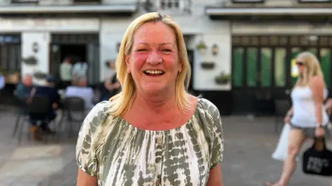 Trish is outdoors in front of a white pub building with black trim, smiling at the camera; blurred people sit and walk behind on a patio area.