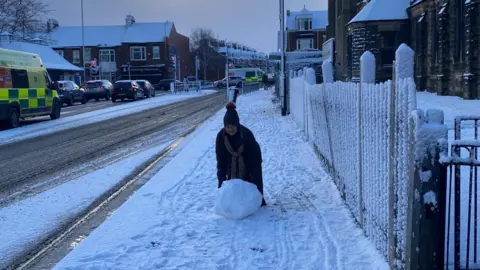 Pallion Action Group and Kayll Road Library community hub A woman is pushing a snowball, which comes up to her knees, along a pavement. The road is slushy with ice behind her.