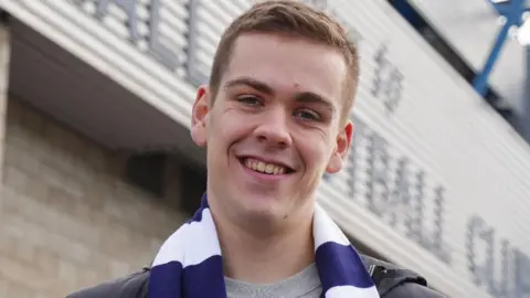 Will Kilgannon in a blue and white striped football scarf. He is standing in front of Millwall Football Club's home ground. 