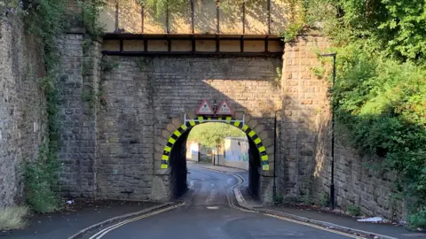 A road narrows as it runs beneath a low railway bridge