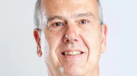 Gloucestershire County Council A photo showing the face of a white man with grey hair. He is stood in front of a white background