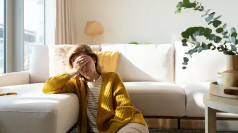 Getty Images A stock image of a woman wearing a yellow cardigan and striped white t-shirt sitting on the floor next to a white corner sofa, with her hand covering her face in apparent discomfort. There is a green potted plant to the right of frame.