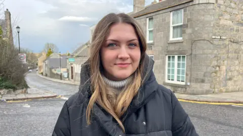 Anastasiia standing in the centre of the shot looking at the camera with the street and residential buildings on the background.