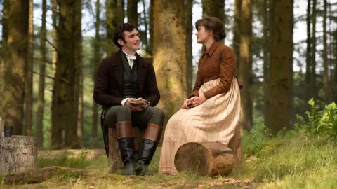 BBC/Bad Wolf/James Pardon Left to right: Mr Hayward (Dónal Finn) wearing a suit in a forest with Mary Bennet (Ella Bruccoleri), who is dressed in a beige and brown dress.