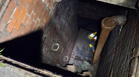 The drain from above. A floodlight appears to be shining down into it. There is a large metal pipe going down the length of a dark brick wall, and other metal bars on the left. There are traffic cones at the bottom of the deep drain.