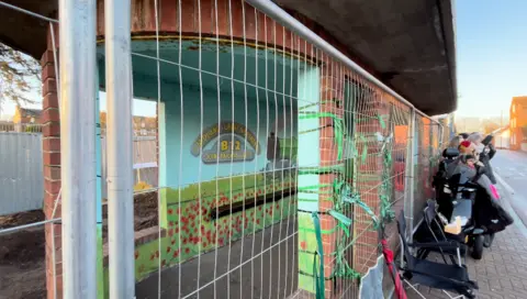 Jack Maclean/BBC Side view of the Sheringham bus shelter - with mural visible through the wire barrier and a group of people next to it on the right hand side