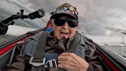 Casey Bukowski onboard a glider plane. He has his mouth open and is wearing a cap, jacket and dark sunglasses. There is a cloudy sky behind him and green fields below.