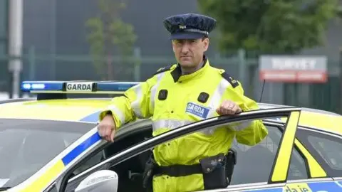 A man wearing an Irish police officer uniform of a yellow hi-vis jacket standing behind the open door of a police car which is stationary 