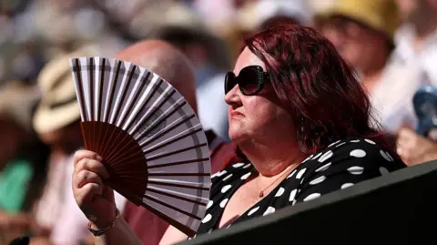 Getty Images A woman with red hair and wearing sunglasses uses a fan to keep her cool while watching the tennis at Wimbledon on 12 July.