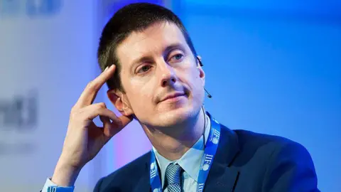 Getty Images The Chair of the Northern Ireland Fiscal Council Sir Robert Chote appears before a blue background. He is wearing a suit and tie as well as a blue lanyard and a microphone ear piece.