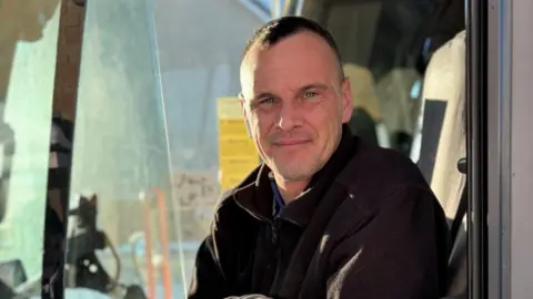 Jim Thow sitting in a tractor cab, looking at the camera. He is wearing a black fleece jacket and has dark hair. 