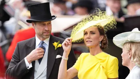 PA Media The Prince and Princess of Wales, William and Catherine, are pictured among crowds at Royal Ascot. Prince William, stood to the left, is wearing a three piece suit with a top hat. Princess Catherine is wearing a yellow dress and hat, the brim of which she is holding with her right hand. 