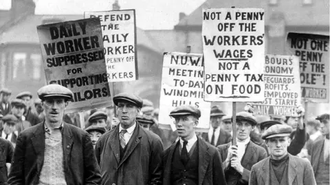 A crowd of men on strike wave placards that carry messages such as "Not a penny off the workers' wages, not a penny tax on food".