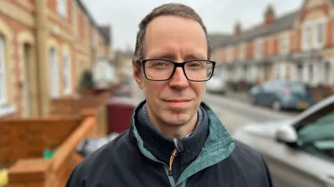 Rob White stands on the pavement on Church Road. He has short grey/black hair and wears black rimmed glasses. He wears a fleece with a coat over it.