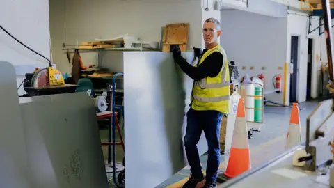Royal British Veterans Enterprise A man wearing safety glasses and a yellow hi-vis jacket is working inside a factory. There are two orange witches hats and he is holding a metal board. 