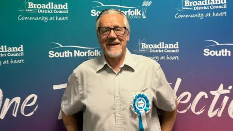 Jimmi Lee is standing in front of a blue and violet panel which has the branding of Broadland District Council and South Norfolk District Council printed on it. He's wearing a light coloured shirt, blue and white Reform rosette and dark rimmed glasses.