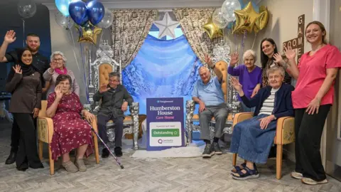 Humberston House Residents and staff are pictured either side of a cardboard sign, with five people on each side. The sign reads Humberston House care home inspected and rated as outstanding. There are blue, white and gold balloons behind them and they are all waving at the camera.