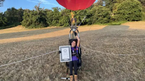 Jo Bailey, wearing brown boots, black leggings and a purple t-shirt. She is sitting in a harness strapped to a hot air balloon, hovering slightly about the ground. She is at the bottom of a sloped hill in a field covered in dry grass, trees can be seen on the horizon. Bailey is holding a framed copy of the Guinness World Record with her right arm and smiling at the camera.