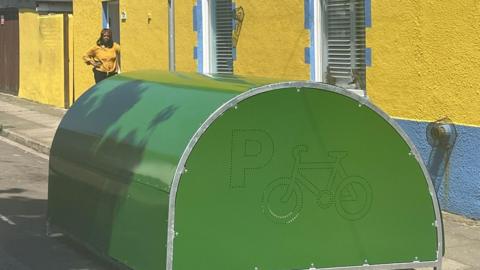Bright green bike hangar on the road in a street in portsmouth with Veloris Ballingall stood outside her bright yellow and blue home in Portsmouth