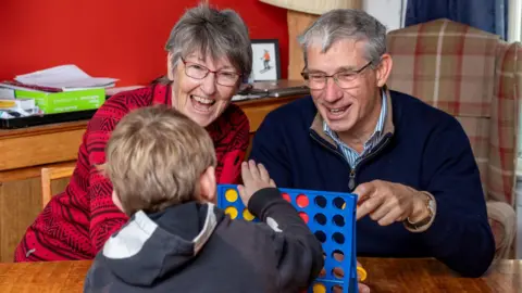 North Yorkshire Council Judith and Martin Ellis sit at a dining table, playing the game Connect Four with a young child with their back to the camera. Judith has short grey hair and wears a red-framed glasses and a red jumper with black patterns. Martin has short grey hair and wears black-framed glasses, a blue and white striped shirt and a blue jumper.