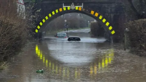 Severe flooding under Lockerley Rail bridge. Two cars are almost entirely submerged under the water. A green food waste caddy is seen floating by. 