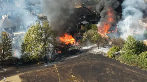 An aerial view of a fire engulfing gardens, with trees either side and house nearby. There is a scorched field in the foreground.