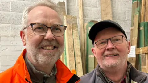 Wolverhampton Wood Recycling Eimert is smiling at the camera as he stands with a volunteers in an orange jacket in the workshop with wood panels visible behind them. The volunteer has glasses on and is also smiling in the headshot