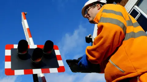 A man wearing an orange hi-vis fleece is to the right of the photo. He also has a white protective hat on, safety glasses and blue gloves. He's holding a electronic tablet and is looking up to a level crossing warning light, which has a red and white striped border, and three coloured lamps. 
