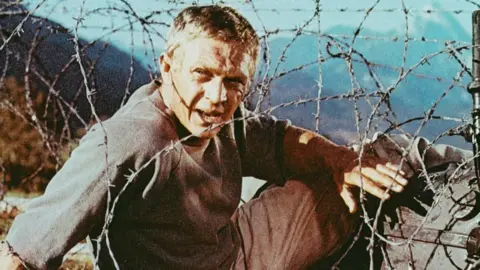 Getty Images Steve McQueen in a publicity shot for The Great Escape, crouching next to a fence of barbed wire.