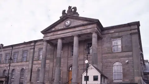 A close-up of Omagh courthouse on a cloudy day.  The large, grey stone building has arched Georgian windows and a Palladian portico with a large clock. 